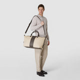 Frontal view: A man carries the TRAVEL BAG IN CANVAS AND MOSAICO over his shoulder; natural-colored canvas with an espresso-colored mosaico stripe and cashmere leather details against a neutral background.