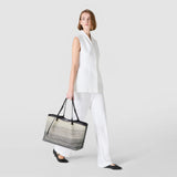 Frontal view — woman holds the SECRET TOTE BAG IN CANVAS in her right hand; embroidered white-and-black gradient canvas with black leather handles, against a neutral light gray background. Text: SERAPIAN