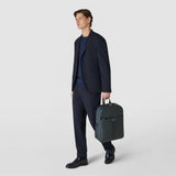 Frontal view: BACKPACK IN CACHEMIRE LEATHER AND MOSAICO is worn by a man in a suit; he holds it by the top handle beside his leg against a light background.