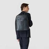 Rear view: BACKPACK IN CACHEMIRE LEATHER AND MOSAICO worn by a man, shows navy blue grained leather and a central Mosaico stripe, elegant business outfit, neutral background. Text: "SERAPIAN"