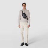 Frontal view: SLING BACKPACK IN CACHEMIRE LEATHER AND MOSAICO, worn diagonally across the chest, black grained leather, Mosaico detail on the strap, model against a light neutral background.