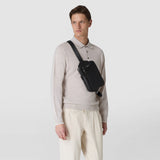 Frontal view: SLING BACKPACK IN CACHEMIRE LEATHER AND MOSAICO, worn diagonally across the chest by a man, black rectangular Cachemire leather compartment with embossed Mosaico shoulder strap and visible 'SERAPIAN' inscription, light background.