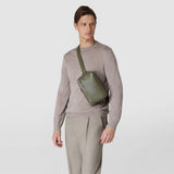 Frontal view: Man wears SLING BACKPACK IN CACHEMIRE LEATHER AND MOSAICO across his chest, leaf-green Cachemire leather, Mosaico-embossed shoulder strap, neutral light studio background.