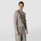 Frontal view: Man wears SLING BACKPACK IN CACHEMIRE LEATHER AND MOSAICO across his chest, olive-green rectangular leather with a Mosaico-embossed strap, neutral background. Text: 'SERAPIAN'.