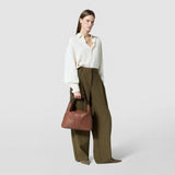 Front view: A woman holds the ANI SHOULDER BAG IN CACHEMIRE LEATHER AND MOSAICO in tobacco brown with Mosaico details in her right hand, elegantly dressed.