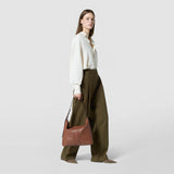 Front view: A woman carries the ANI SHOULDER BAG IN CACHEMIRE LEATHER AND MOSAICO in tobacco brown on her shoulder. The bag features Mosaico details and an adjustable shoulder strap.