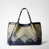 Frontal view: SECRET TOTE BAG IN DENIM AND RAFFIA stands against a neutral background, shows navy-blue jacquard denim and raffia texture, dark-blue leather handles; visible 'SERAPIAN MILANO' lettering.