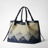 Frontal view: SECRET TOTE BAG IN DENIM AND RAFFIA, presented standing on a neutral background, dark blue leather handles upright, jacquard denim with geometric raffia pattern; visible 'SERAPIAN MILANO' lettering.
