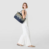 Side view: Model wears SECRET TOTE BAG IN DENIM AND RAFFIA casually over her shoulder, navy-blue patterned large tote, against a light neutral background.