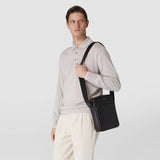 Frontal view: Man wears NORTH SOUTH CROSSBODY BAG IN EVOLUZIONE LEATHER casually over his shoulder; the flat black leather bag rests at his side against a plain light background.
