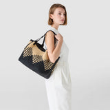 Frontal view: woman wearing SECRET TOTE BAG IN RAFFIA over her shoulder, natural and black crocheted raffia in a Mosaico pattern, casual look against a light background.