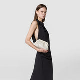 Front view: A woman holds the MINI SECRET CLUTCH BAG IN MOSAICO MICROCHAIN in front of her black dress; the cream-colored bag features a woven, raised mosaic pattern.
