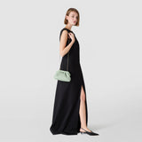 Side view: Model wears MINI SECRET CLUTCH BAG IN RIBBON MOSAICO on a slim gold chain over her shoulder, Aqua Mosaico with three-dimensional texture, black maxi dress, neutral background.