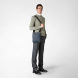 Frontal view — Man wearing FLAT CROSSBODY BAG IN STEPAN slung across his shoulder; ocean-blue textured Stepan material, black adjustable strap, compact rectangular shape, neutral studio background.