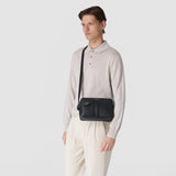 CAMERA CROSSBODY BAG IN STEPAN worn across the body, black embossed Stepan coated canvas with front leather bands, frontal view modeled against a light neutral background. Plaque reads "SERAPIAN".